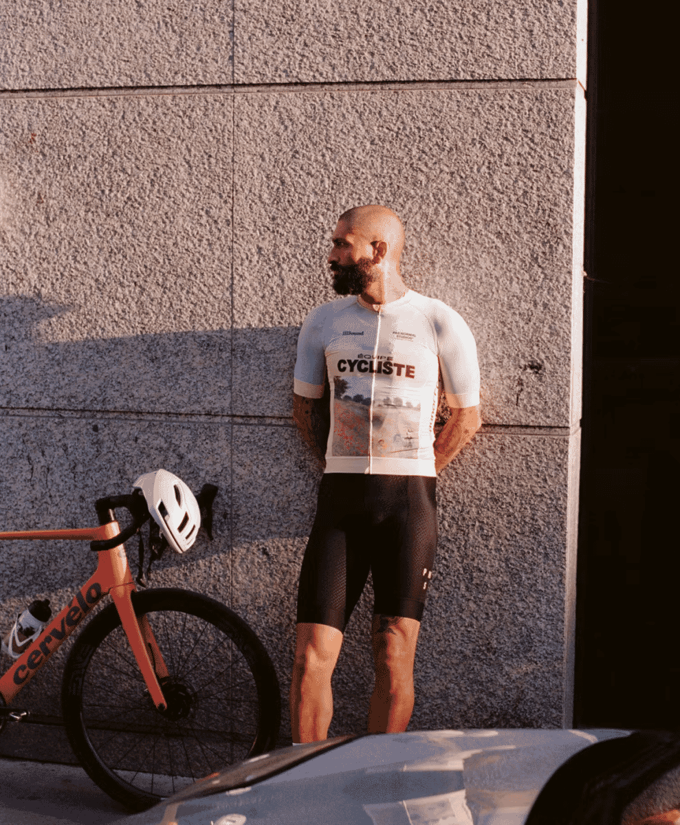 A man in a white 'ÉQUIPE CYCLISTE' jersey standing beside an orange road bike against a textured urban wall.