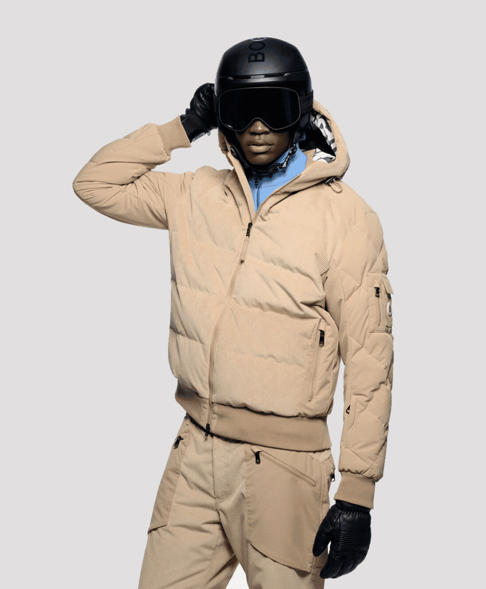 A model in a beige corduroy ski jacket and matching pants with a black helmet and goggles.