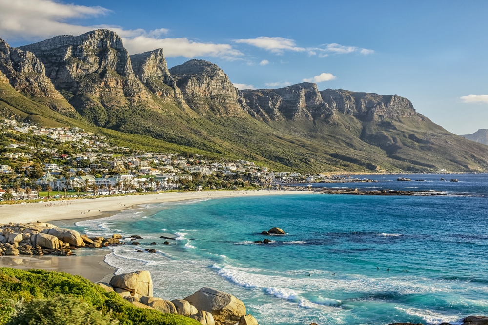 A wide coastal view of a sandy beach and turquoise ocean nestled against dramatic mountain cliffs.