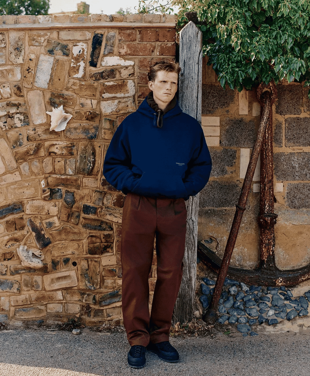 A man in a navy blue hooded sweatshirt standing against a rustic stone wall with maritime decor.
