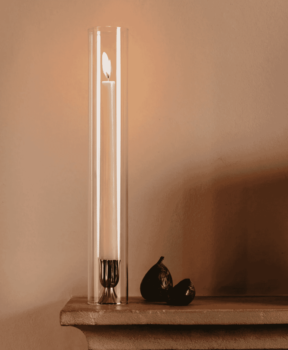 A minimalist still life featuring a lit candle inside a tall glass cylinder beside figs on a stone mantelpiece.
