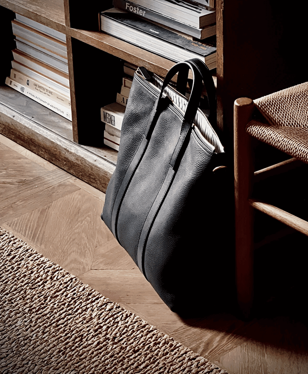 Dark leather tote bag resting on a wooden floor next to architecture books and a bookshelf.