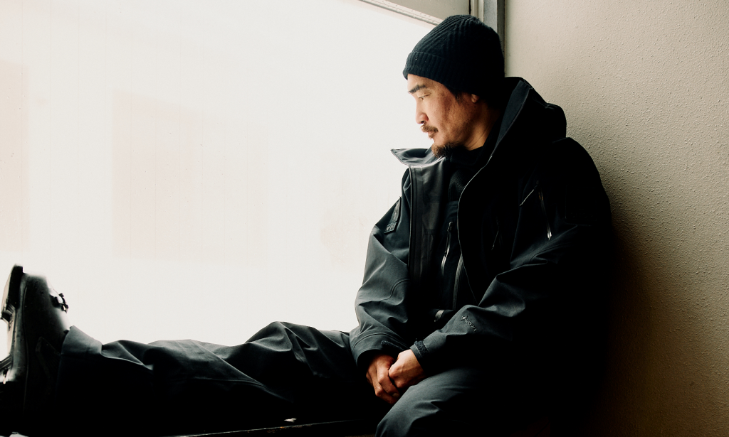 Man in black streetwear-inspired technical outerwear and beanie sitting contemplatively.