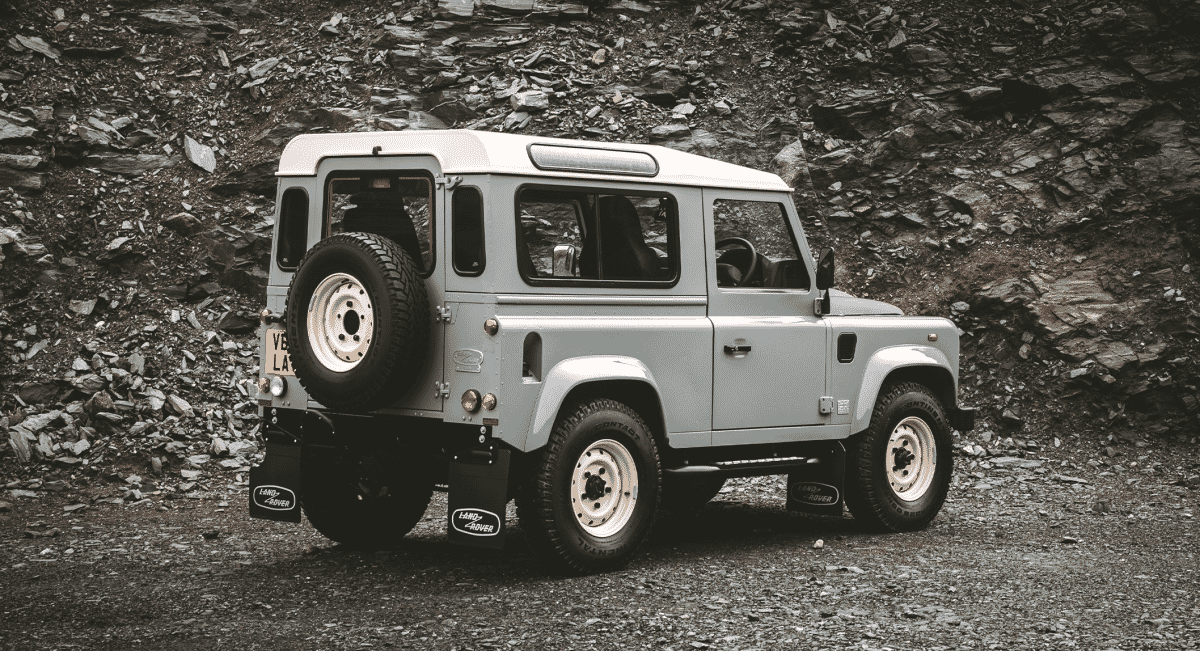 A classic silver Land Rover Defender on rocky terrain showing the rear-mounted spare tire.