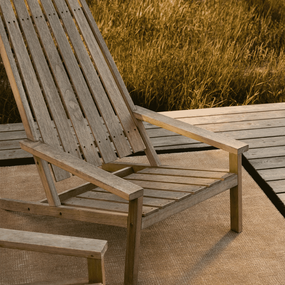 A weathered wooden Adirondack chair sitting on a textured woven rug on a wooden deck.