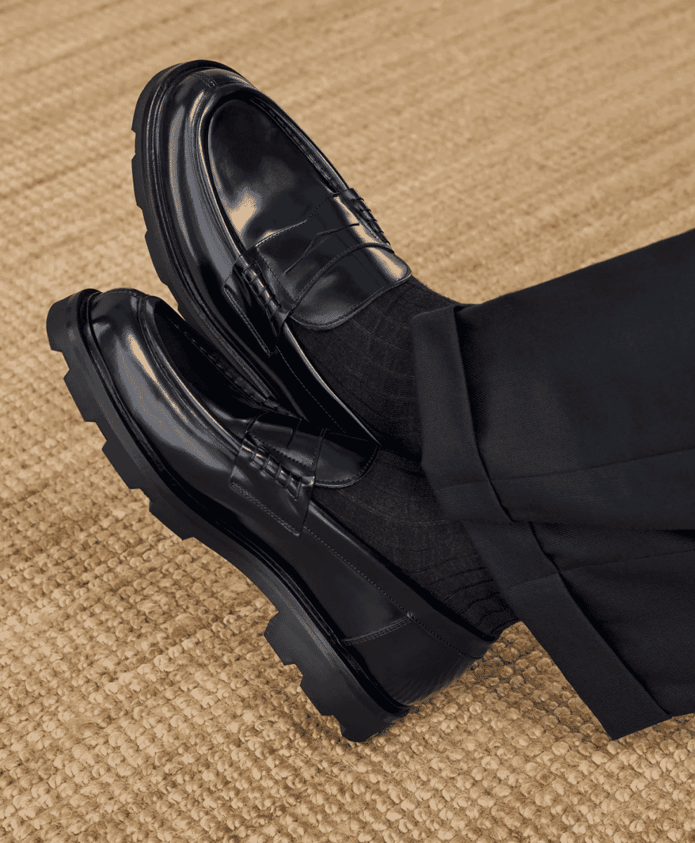 Person wearing glossy black patent leather penny loafers with thick, rugged rubber soles and dark tailored trousers.