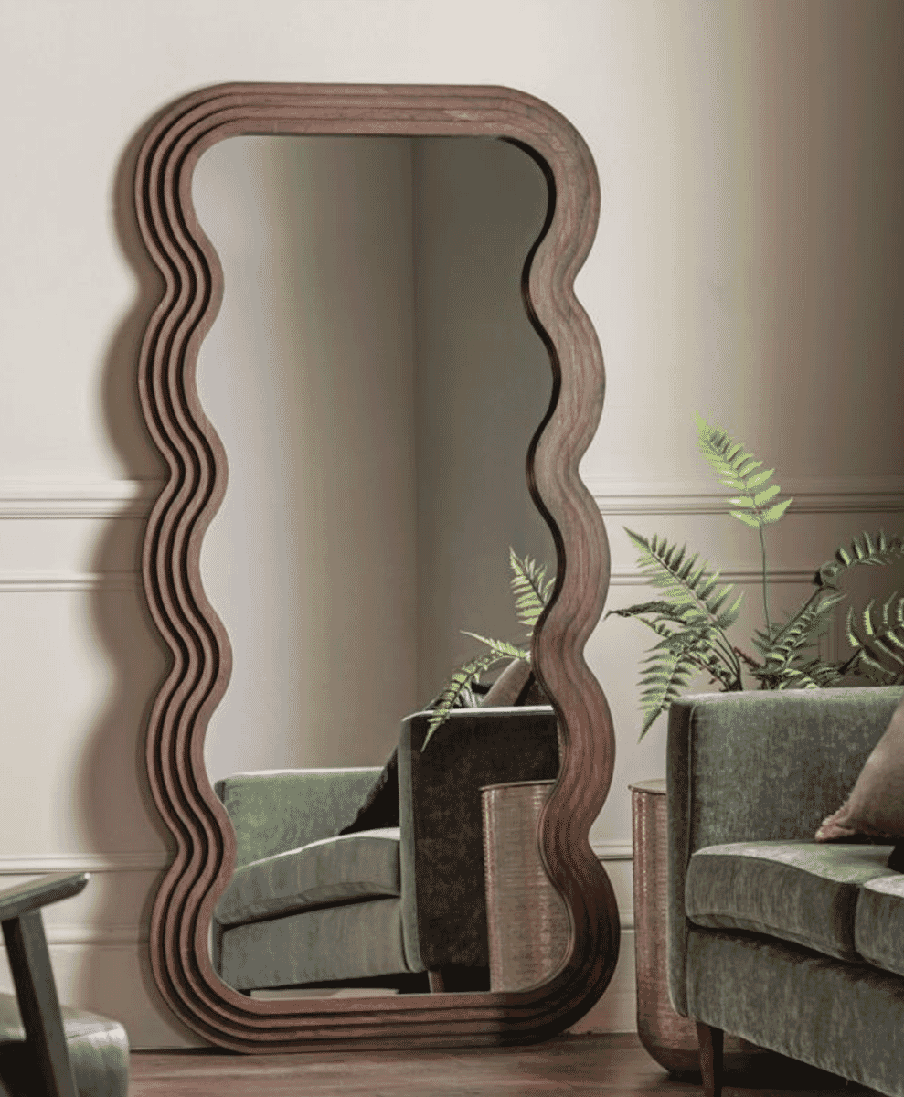 Tall wavy dark wood mirror standing in a living room with green velvet furniture and plants.