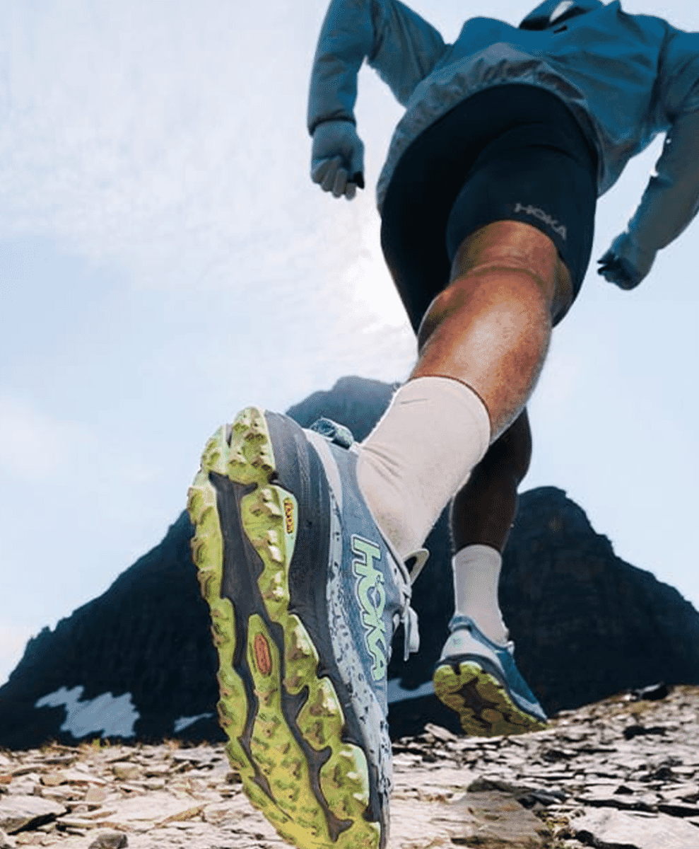 A runner in blue HOKA shoes and black shorts striding uphill on a rocky mountain trail.