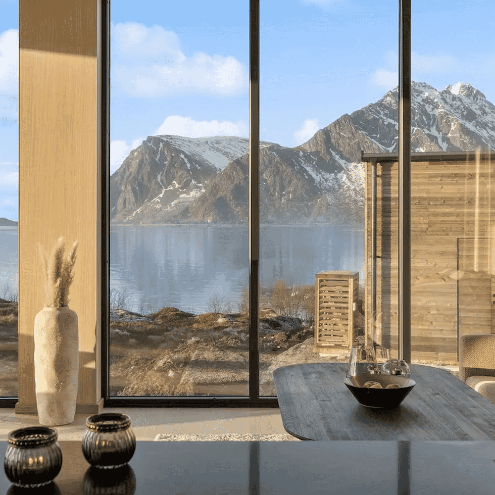 Interior of a modern cabin with wooden furniture and large windows overlooking a snowy mountain fjord.