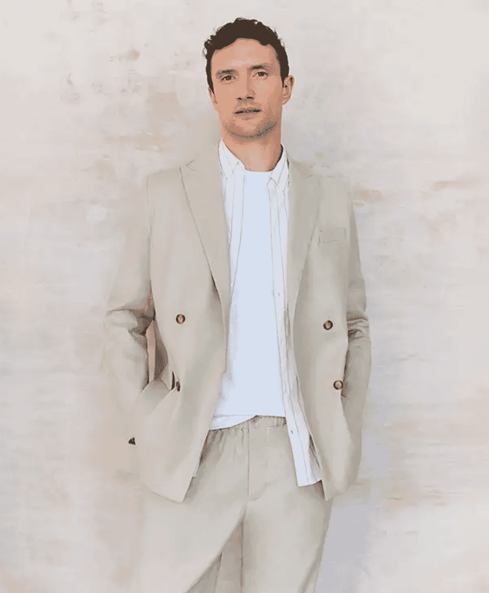 A man modeling a light beige double-breasted blazer against a textured off-white wall.