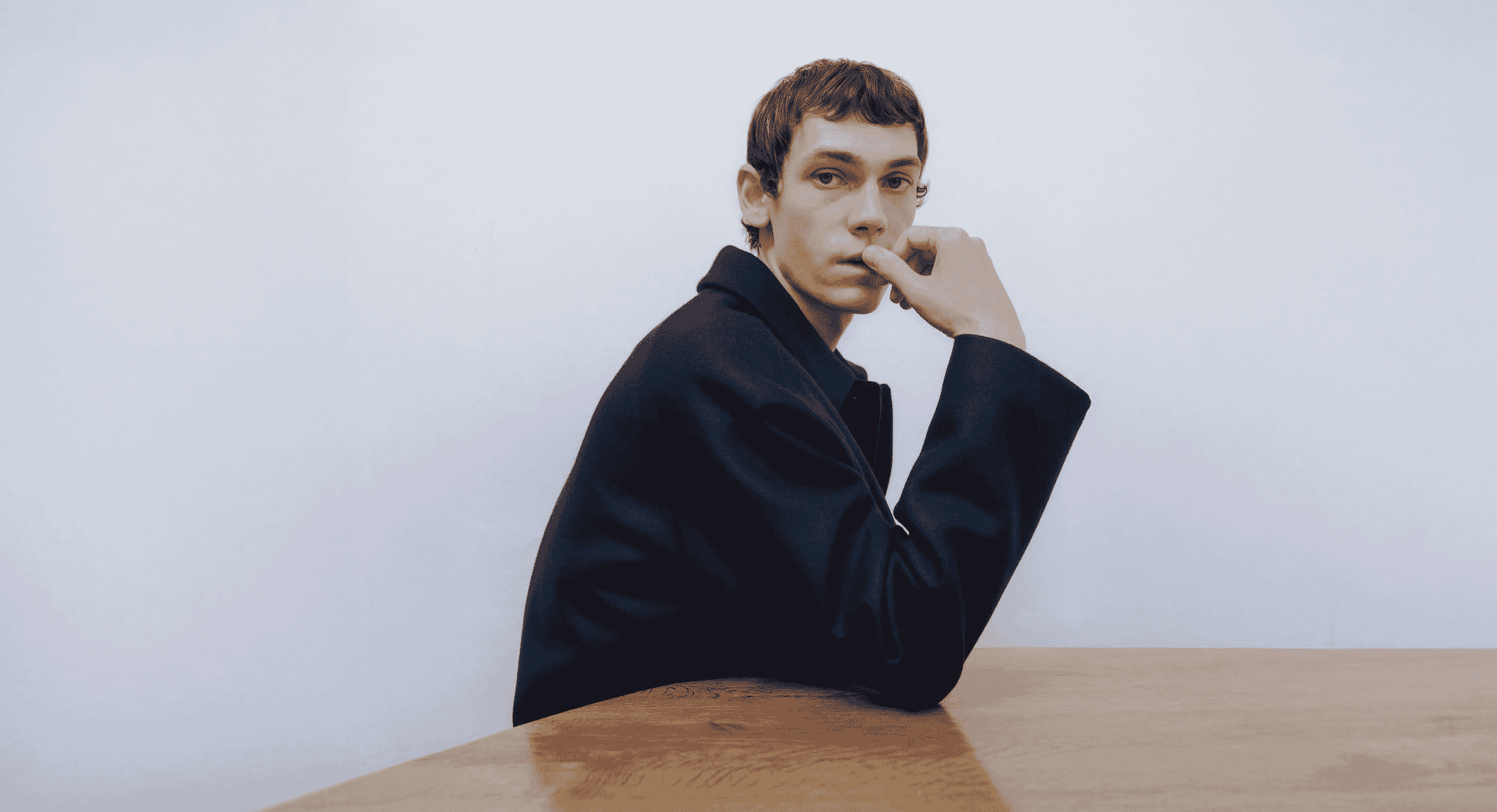 Man in an oversized dark coat posing thoughtfully at a wooden table.