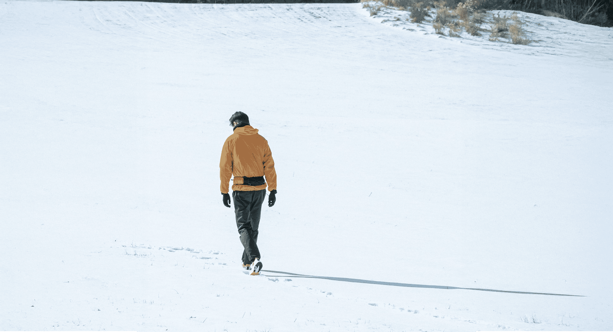 A man in a mustard-yellow hooded jacket and black pants walking across a snow field