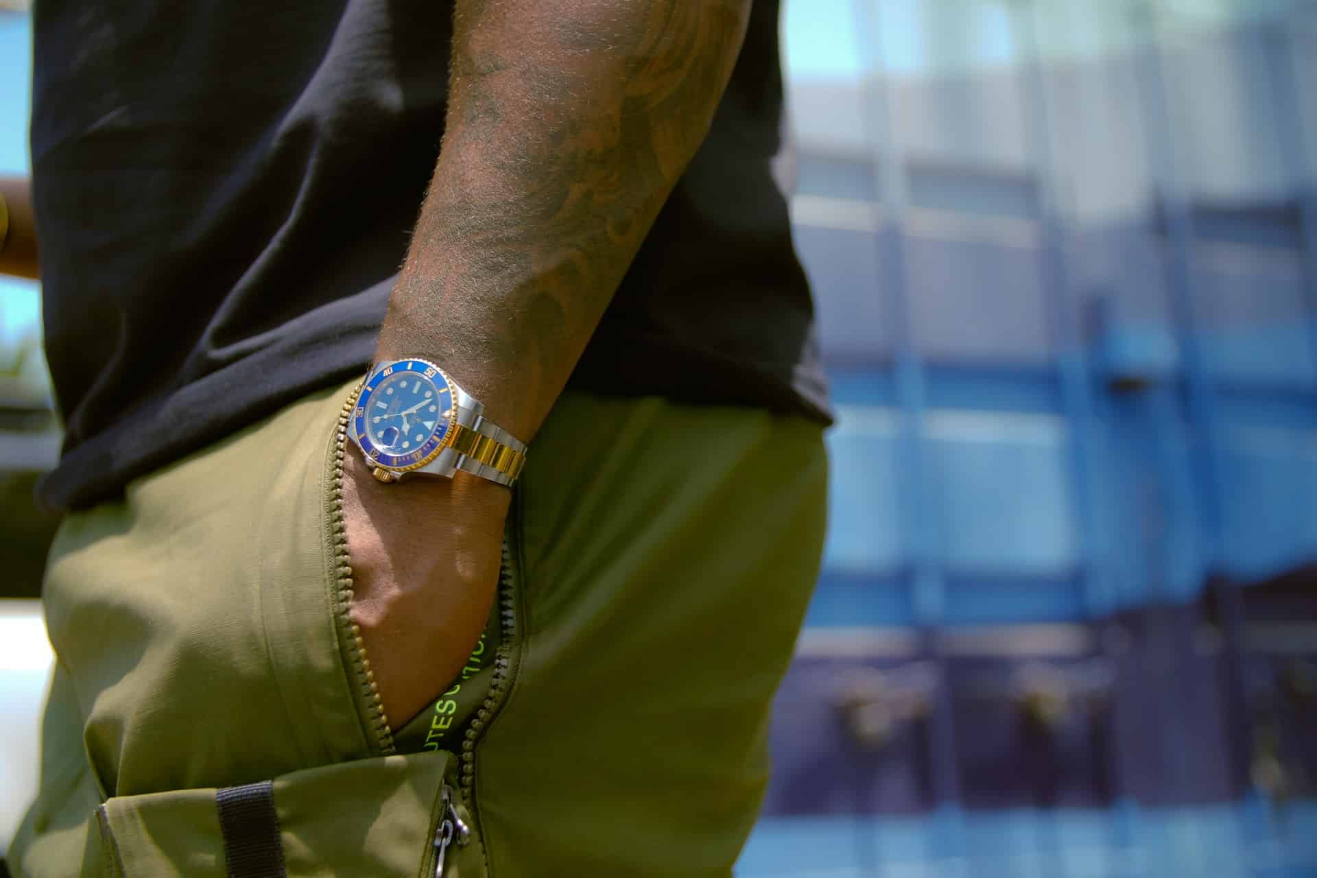 A man with tattoos wearing olive cargo pants and a black t-shirt, showing a blue-dial Rolex Submariner in an urban setting.