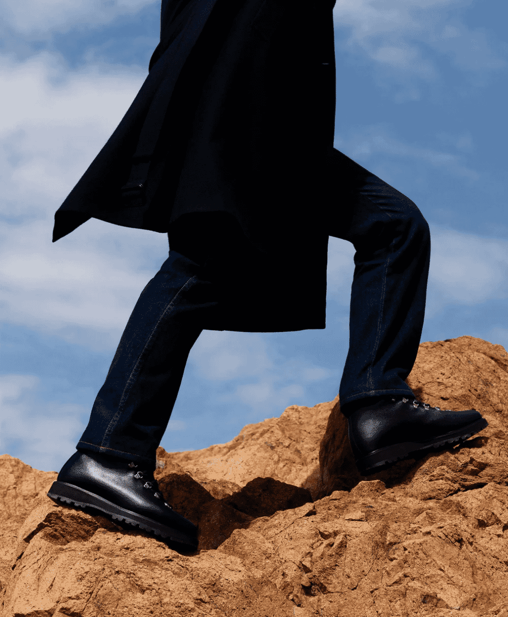 Man in a black coat and rugged black boots walking across rocky terrain.
