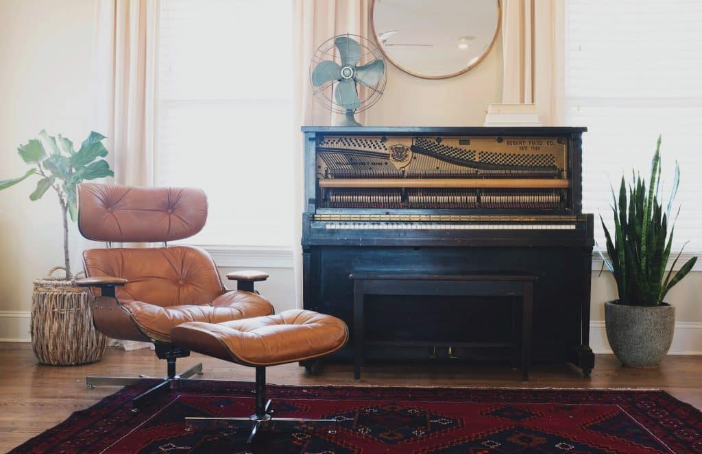 A vintage-inspired room with a brown leather Eames lounge chair, a black upright piano, and a patterned rug.