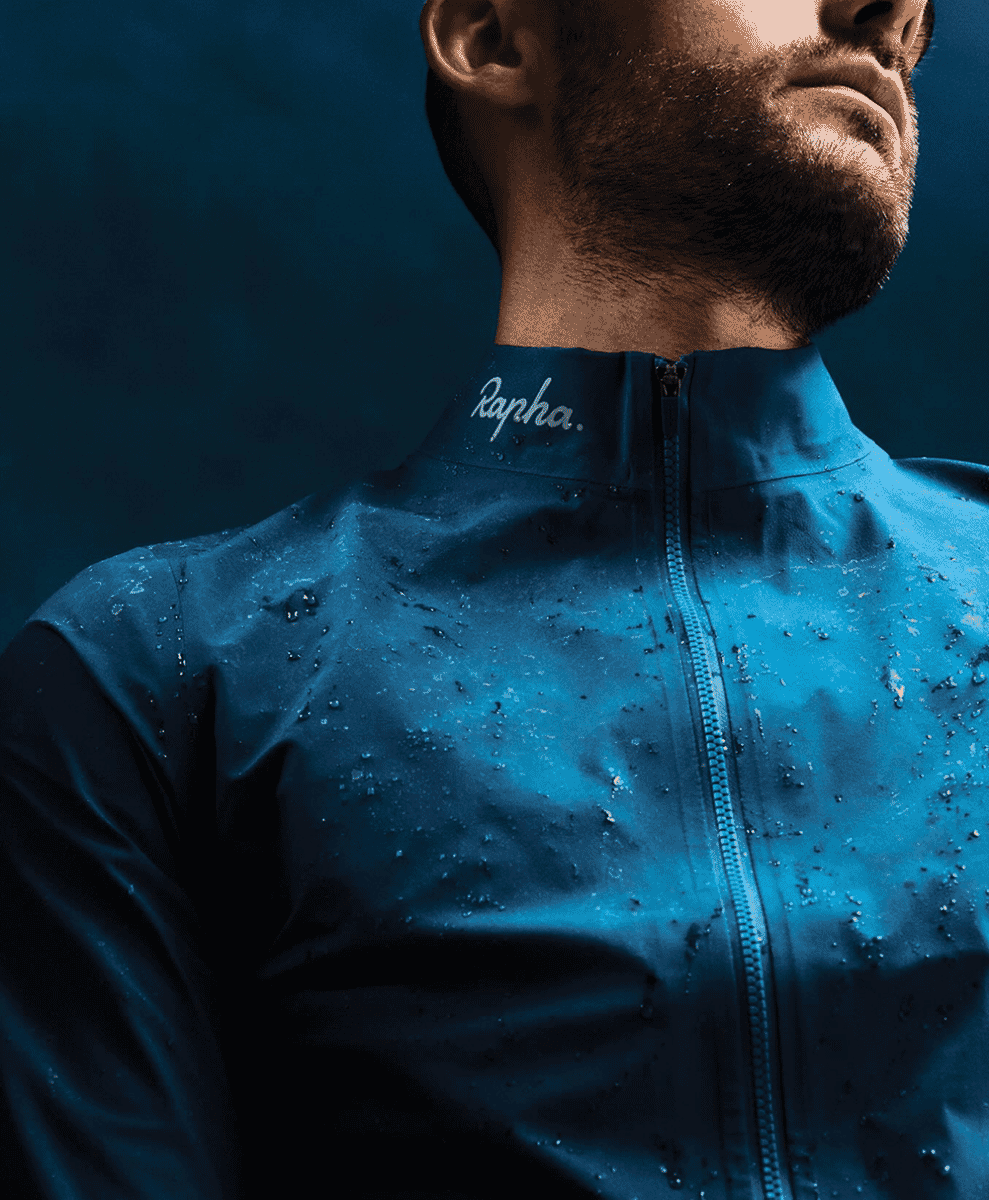 Extreme close-up of water droplets beading on a technical blue waterproof cycling jacket with the Rapha logo.