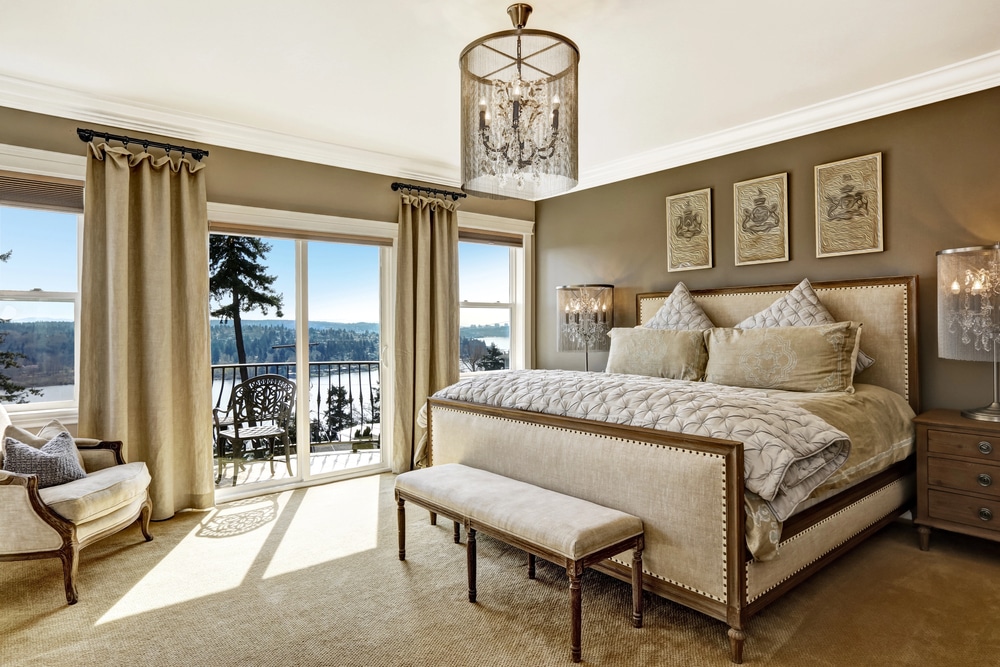 Luxury master bedroom with beige tones, a chandelier, and large windows overlooking a lake.
