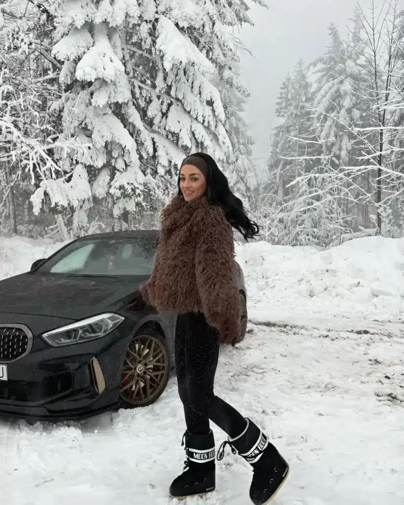 Woman in a fluffy brown coat and black Moon Boots beside a car.
