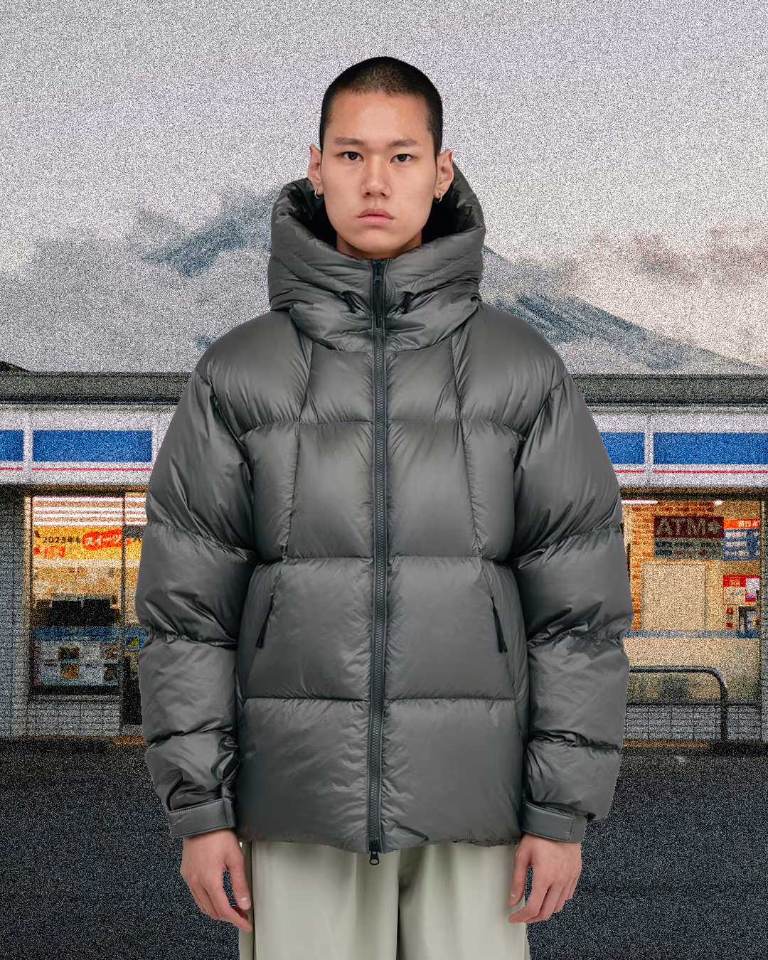 Model in a dark gray puffer jacket against a surreal background of a convenience store and mountains.