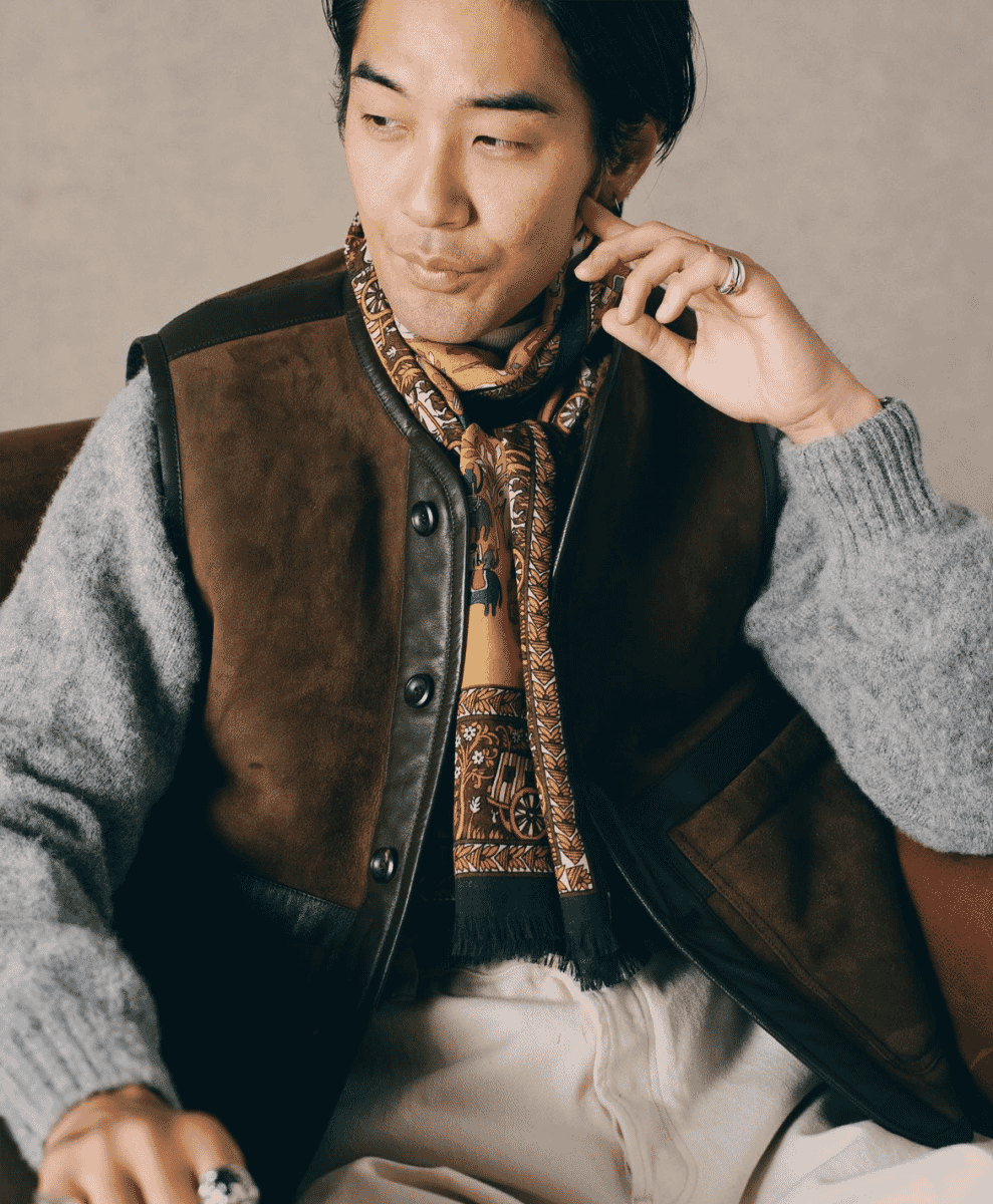 A man wearing a brown suede shearling vest over a grey sweater and scarf.