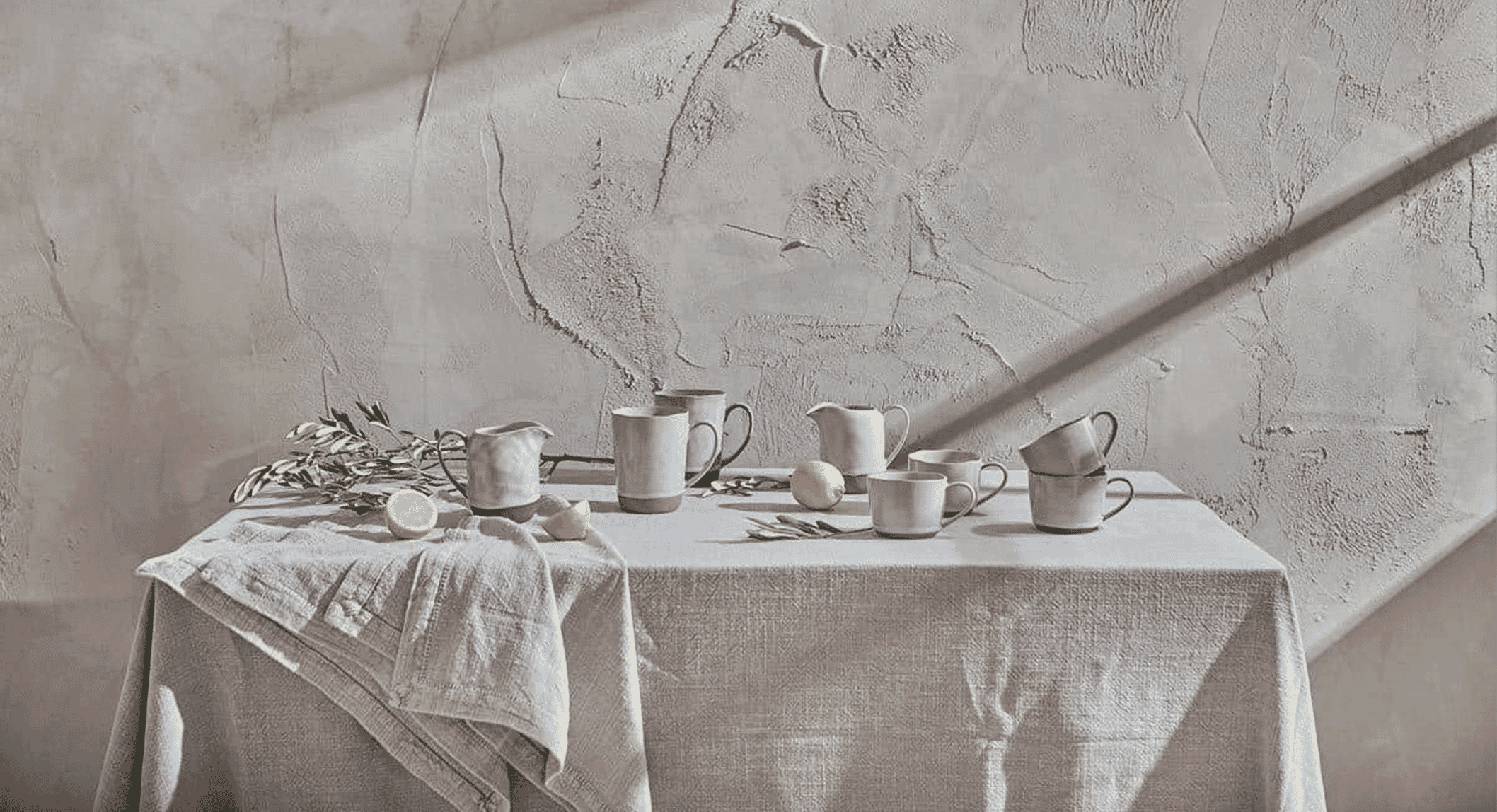 Monochrome still life of ceramic mugs, pitchers, and olive branches on a linen-covered table.