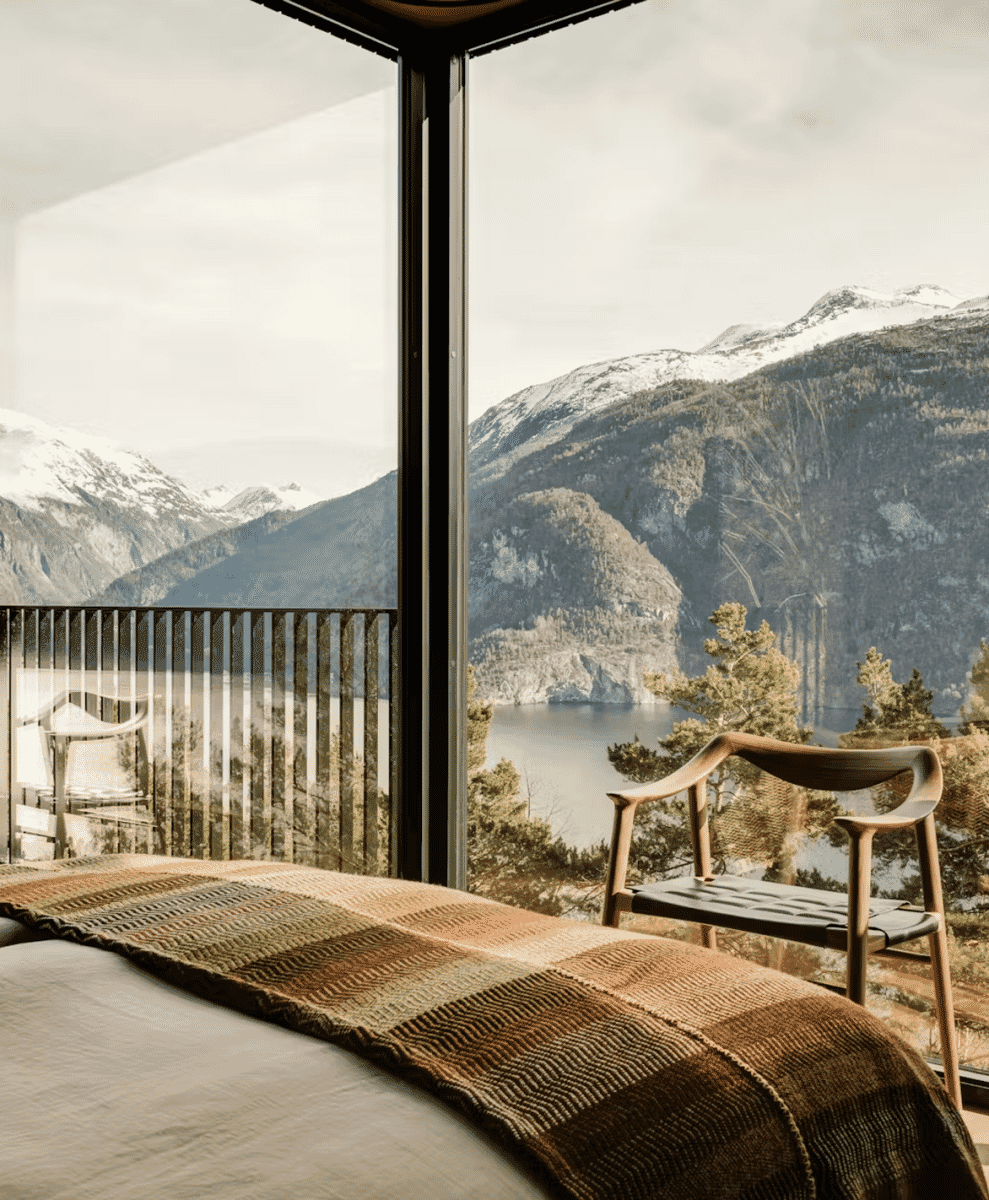 A cozy bedroom with a plaid blanket on a bed overlooking snow-capped mountains through large windows.