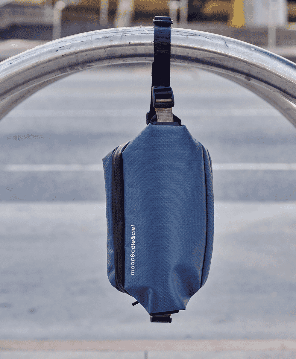 A blue textured sling bag hanging from a metallic bike rack in an urban setting.