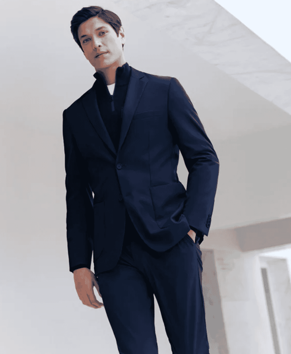 A man in a sleek navy blue suit with a high-necked black top in a minimalist architectural setting.