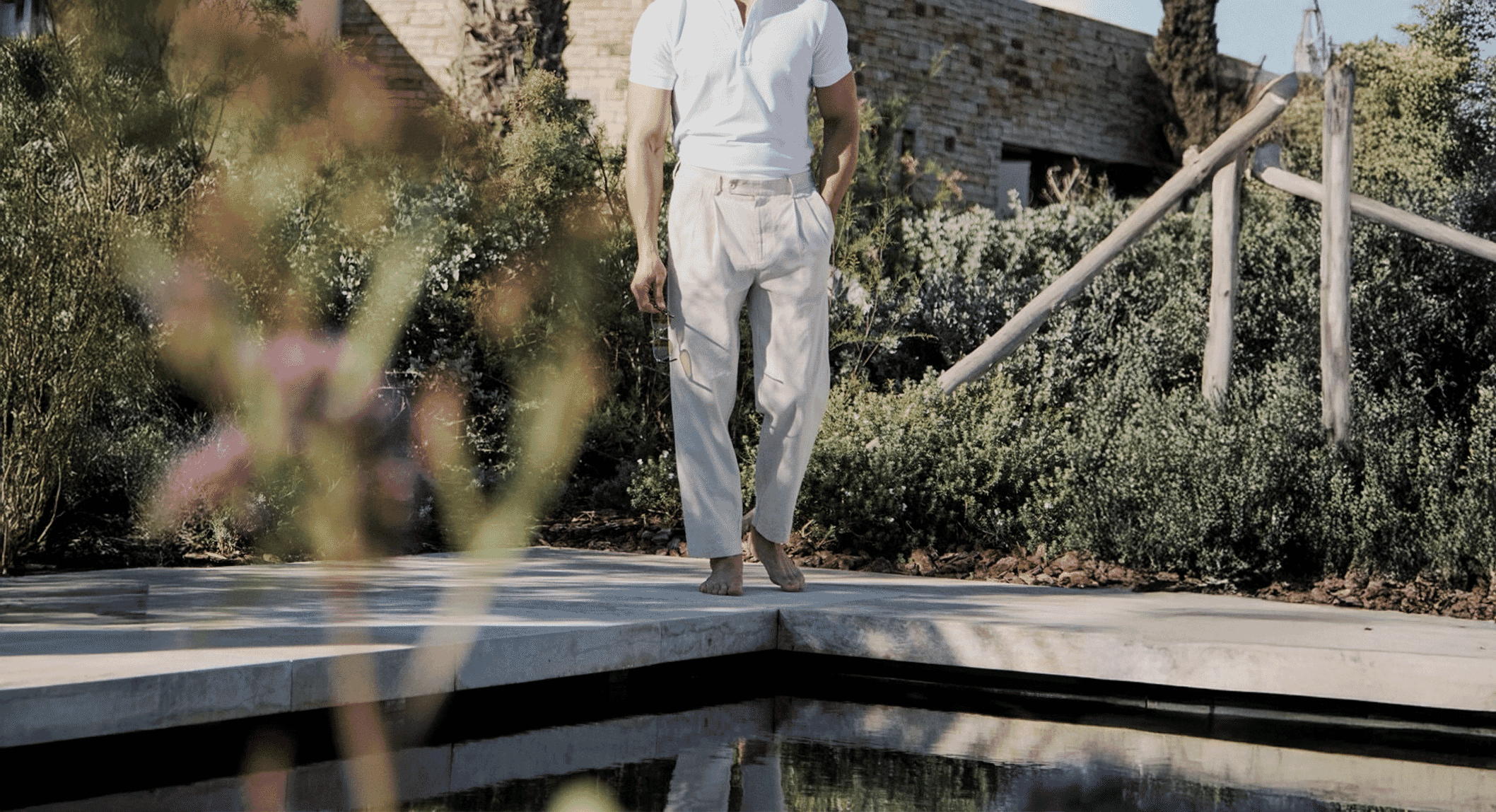 Man in a white polo and light pleated trousers walking barefoot along a sunny poolside patio.