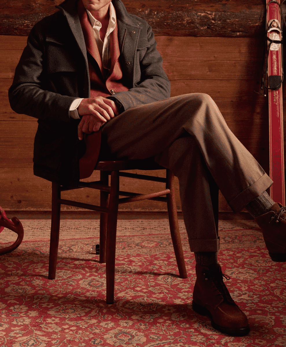 A man in a dark coat and red vest sitting in a rustic cabin setting.