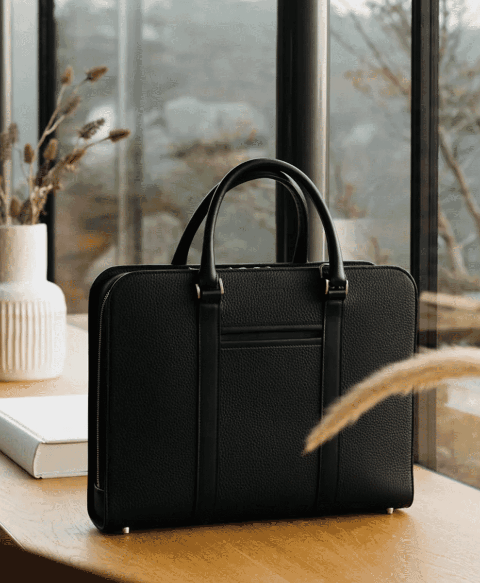 A black pebbled-leather briefcase sitting on a wooden table in a minimalist interior with dried grasses.
