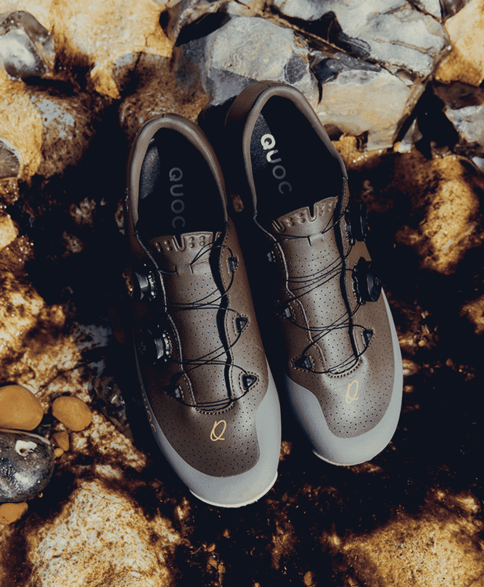 Detail shot of QUOC cycling shoes featuring perforated leather and metallic accents on a wet rocky surface.