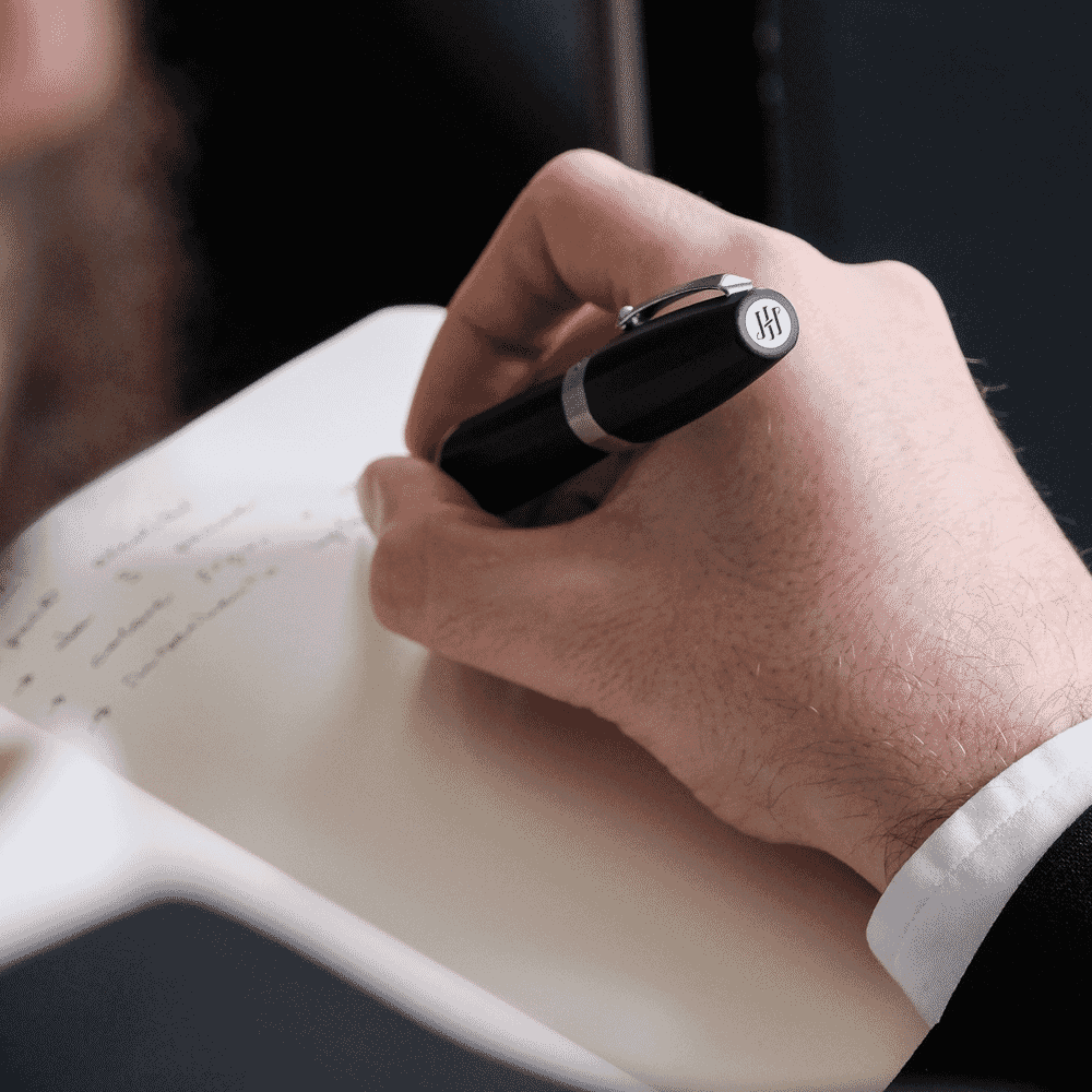 Detail shot of a man's hand writing with a black pen on a notebook over a professional leather surface.