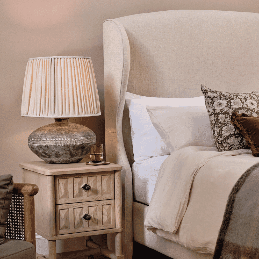 A neutral-toned bedroom featuring a beige wingback bed and a warm lamp on a wooden nightstand.