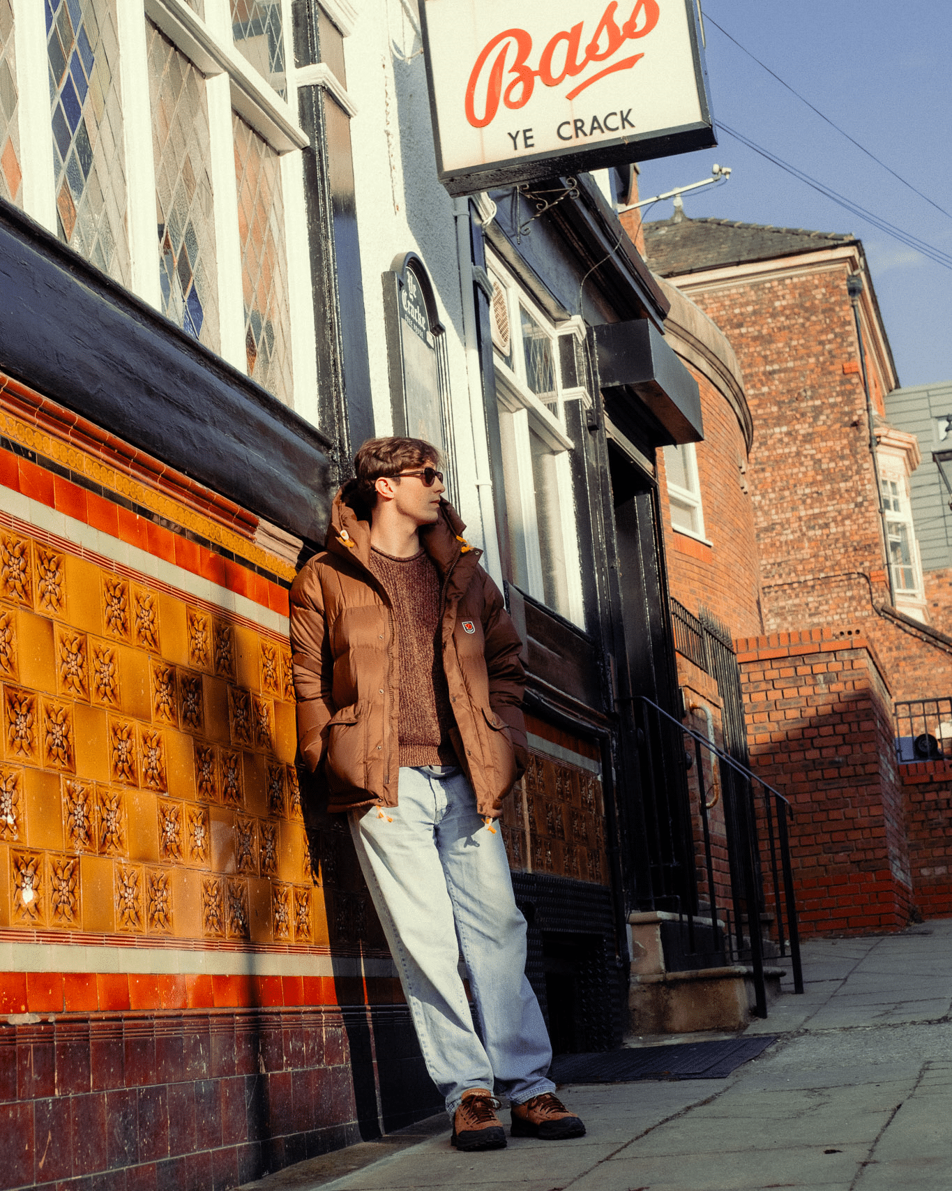 A man in a brown puffer jacket and maroon sweater leaning against a brick wall under a pub sign.