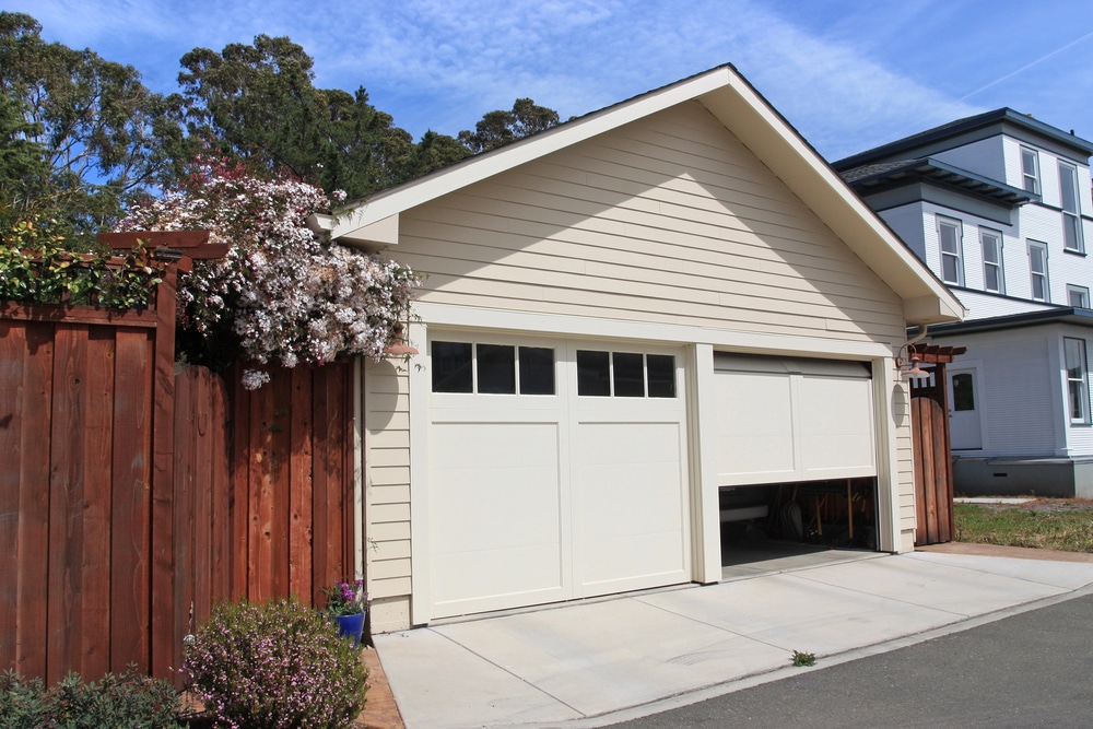 Beige garage doors framed by flowering vines and a wooden fence under a bright blue sky.