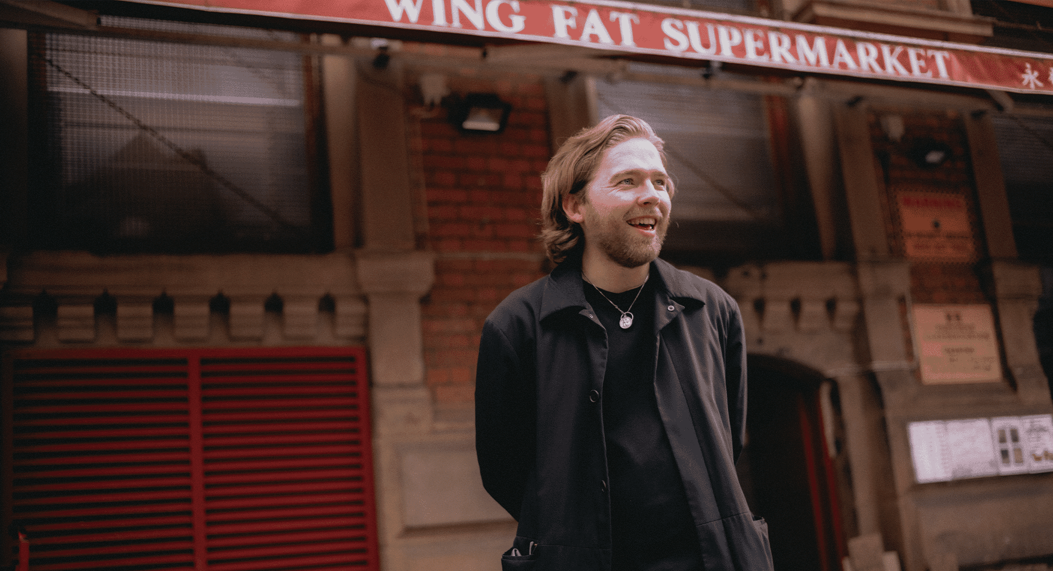 A man in a black coat and necklace standing in front of Wing Fat Supermarket.