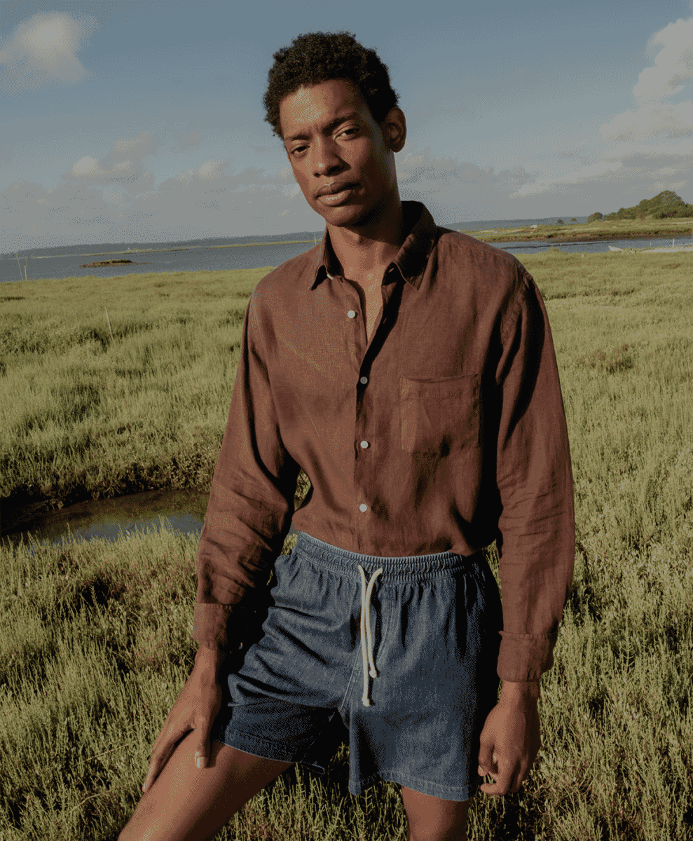 A man models a brown linen button-up shirt and denim shorts in a coastal marshland.