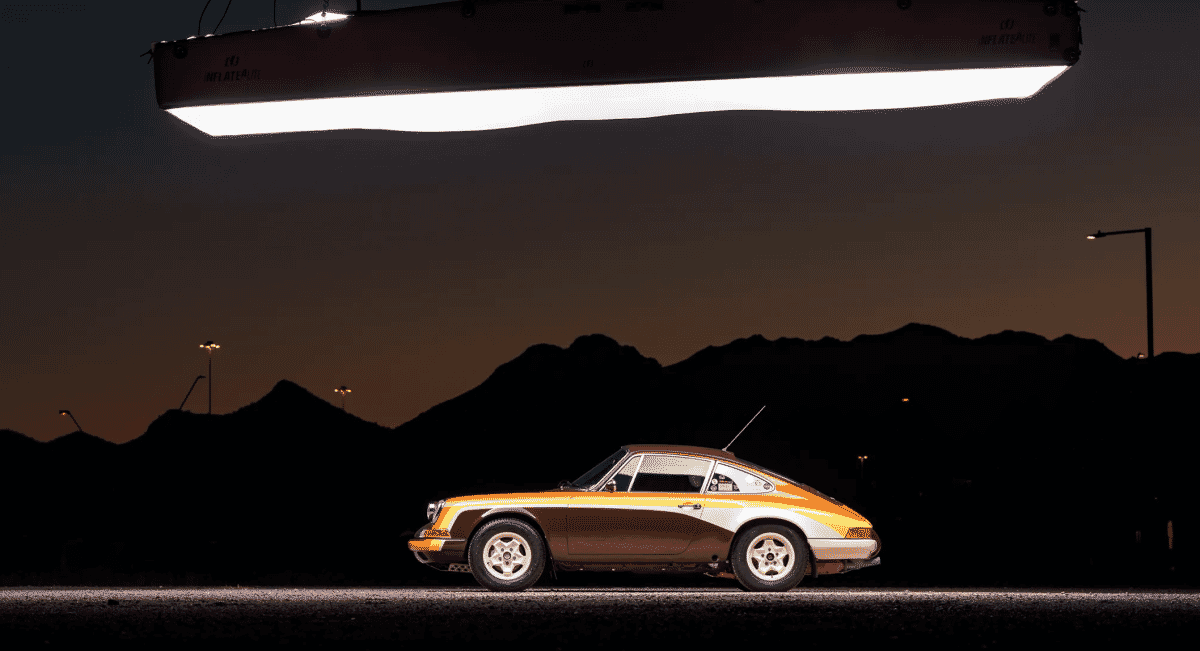 A vintage Porsche sports car with racing livery illuminated under studio lights against a mountain backdrop.
