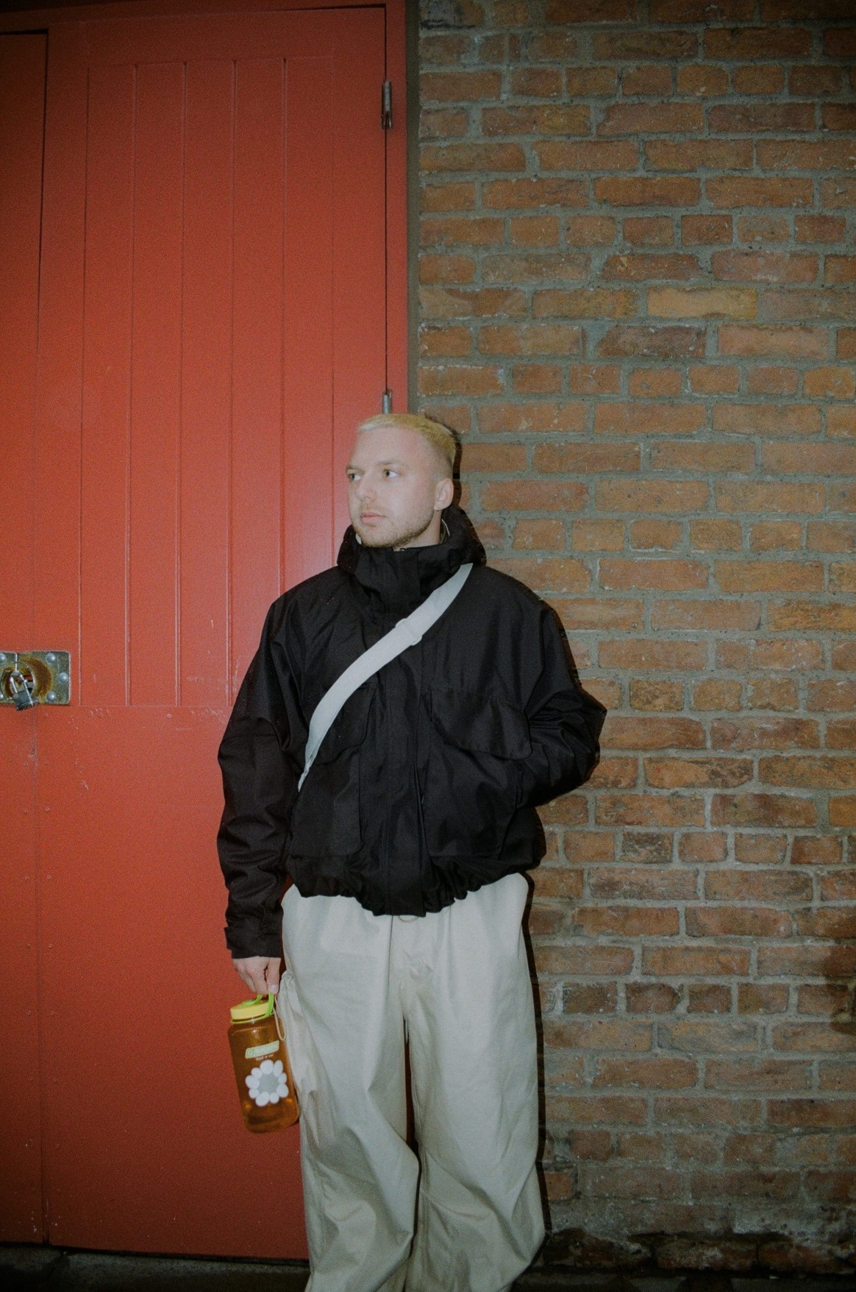 Close-up of a man in an urban setting wearing a black puffer, beige pants, and carrying an orange water bottle.