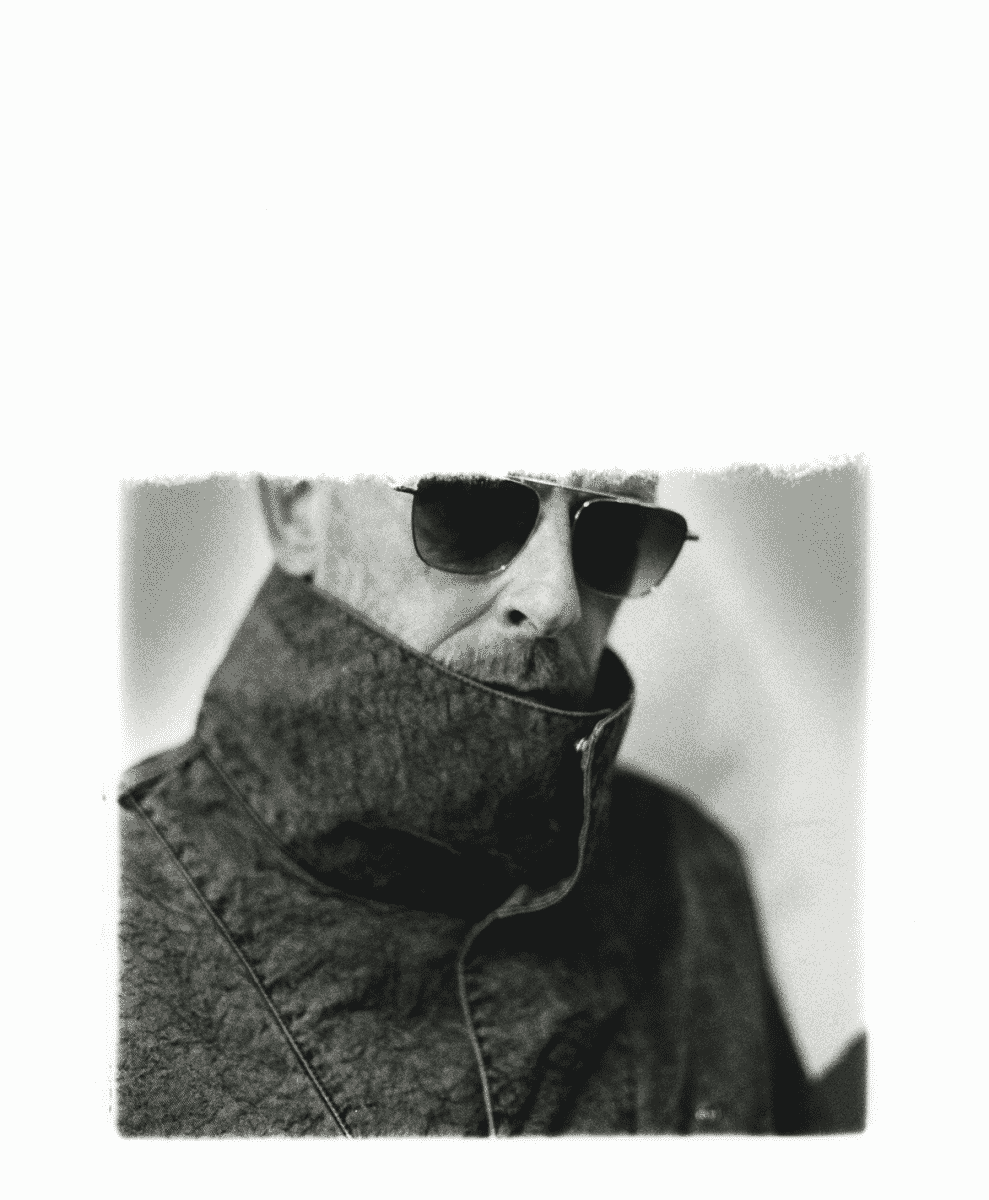 Black-and-white portrait of a man in a high-collared jacket and aviator sunglasses.