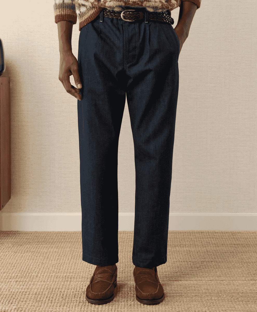 A man modeling dark pleated denim trousers paired with brown loafers and a patterned sweater.