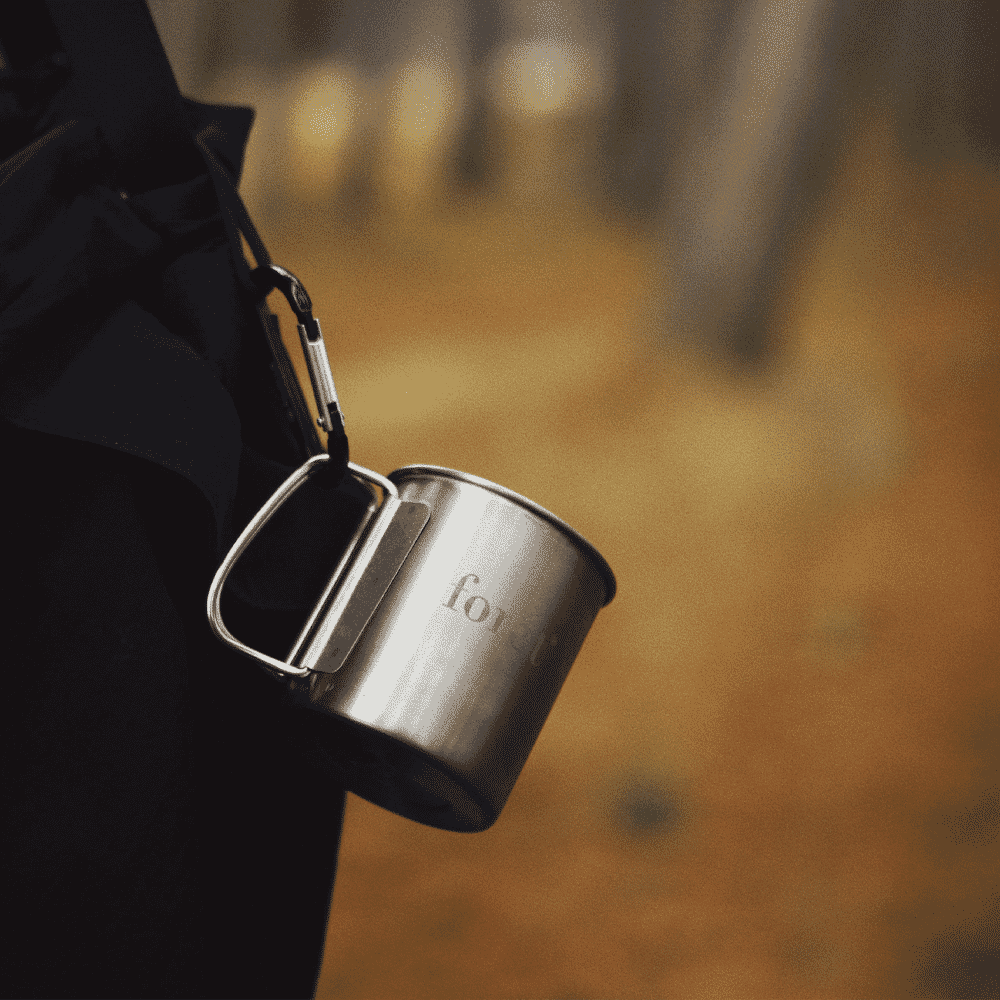 A person in a dark jacket holding a metallic titanium camping mug in an autumn forest.