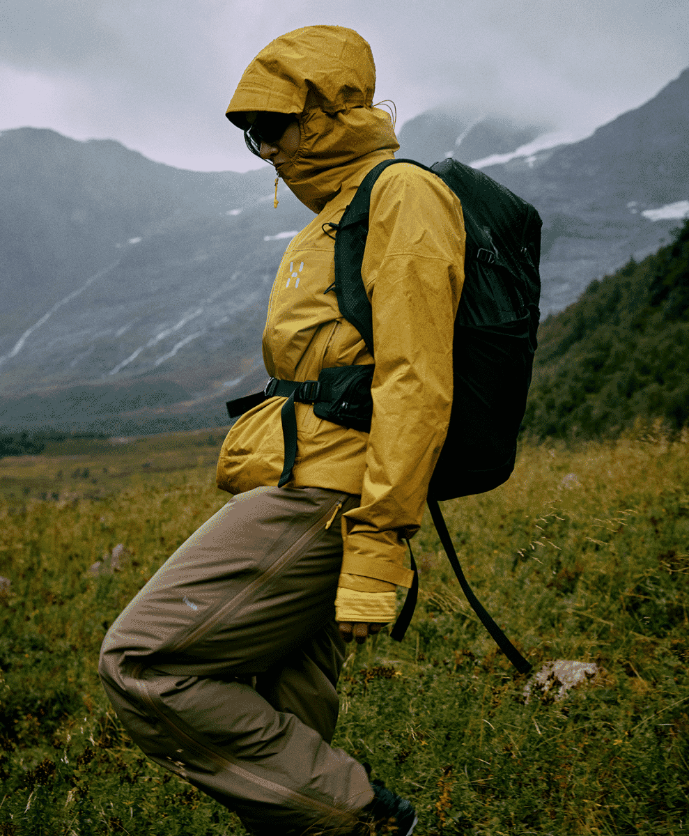 A hiker wearing a mustard-yellow Haglöfs L.I.M jacket and a black backpack on a mountain trail.