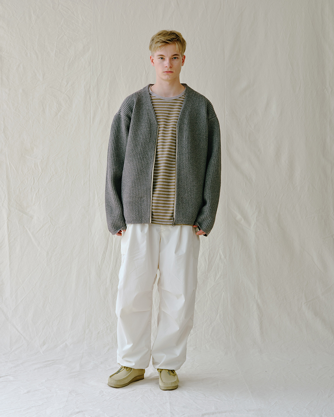 A man wearing a grey textured knit cardigan over a striped tee and white trousers.