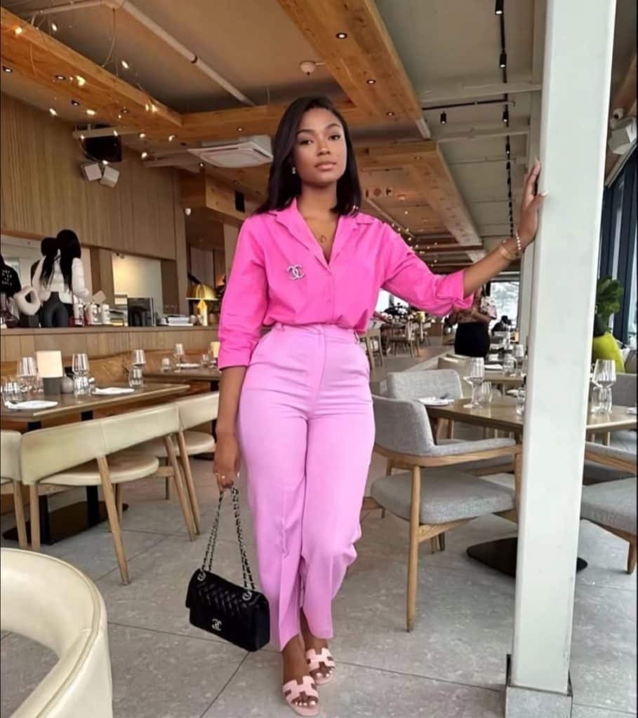 A woman in a tailored pink pantsuit with a Chanel brooch in a modern, stylish interior.