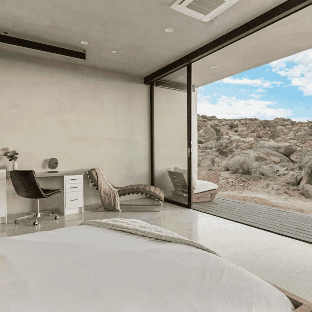 A modern minimalist bedroom with glass doors opening to a bright desert landscape.