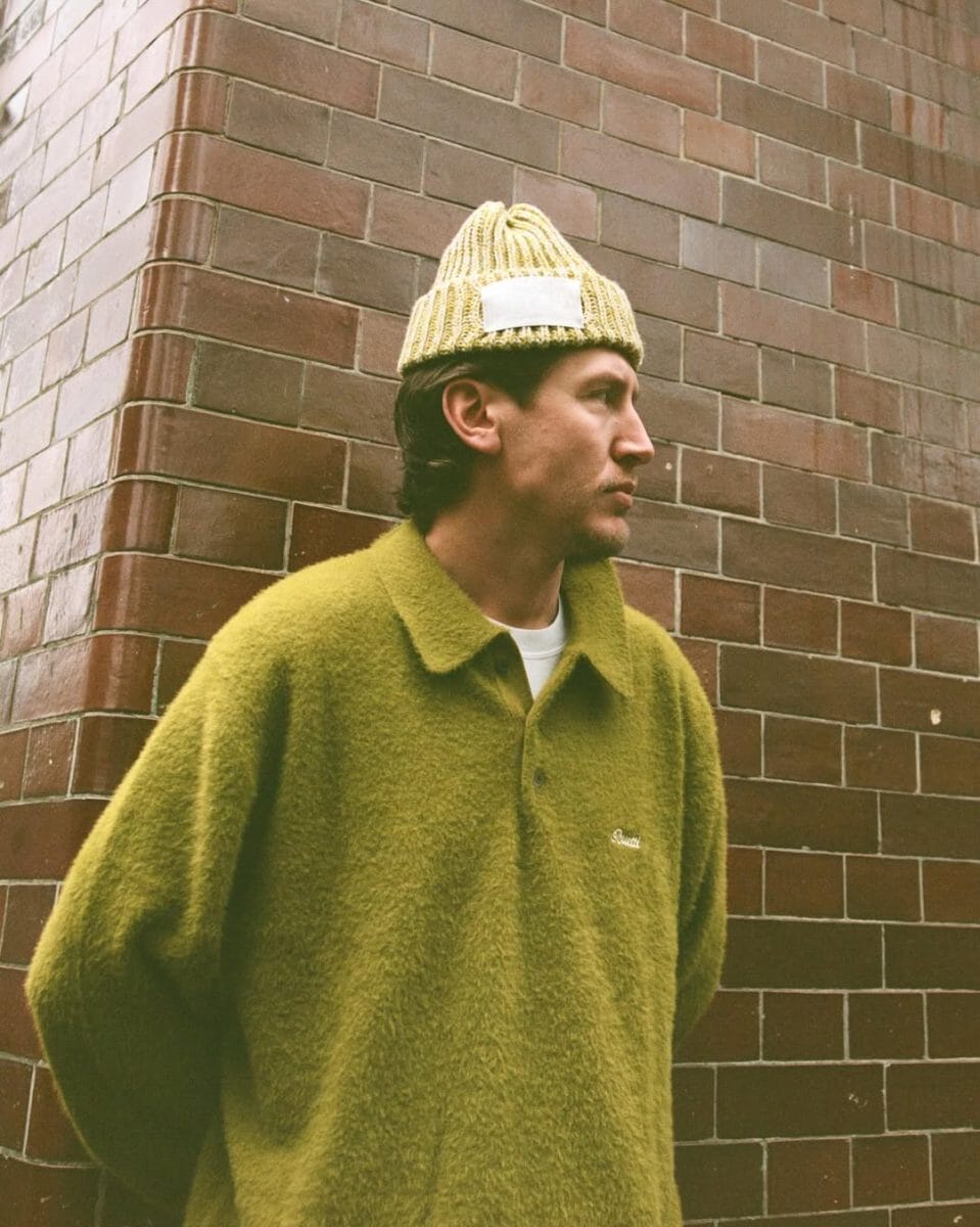 A man in an olive-green knit sweater and beanie standing against a red brick wall.