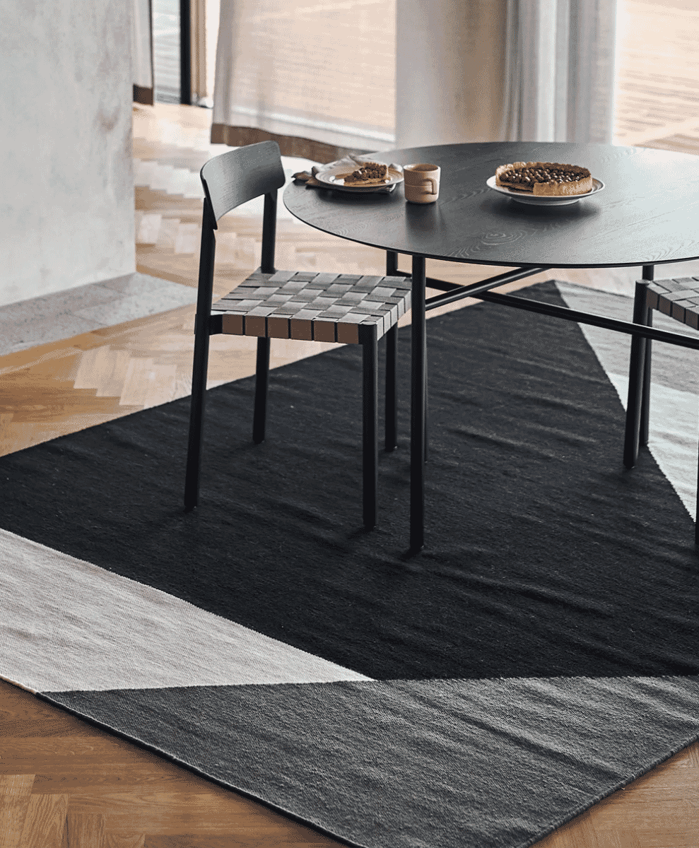 A black round dining table and woven chairs set on a modern geometric low-pile rug.
