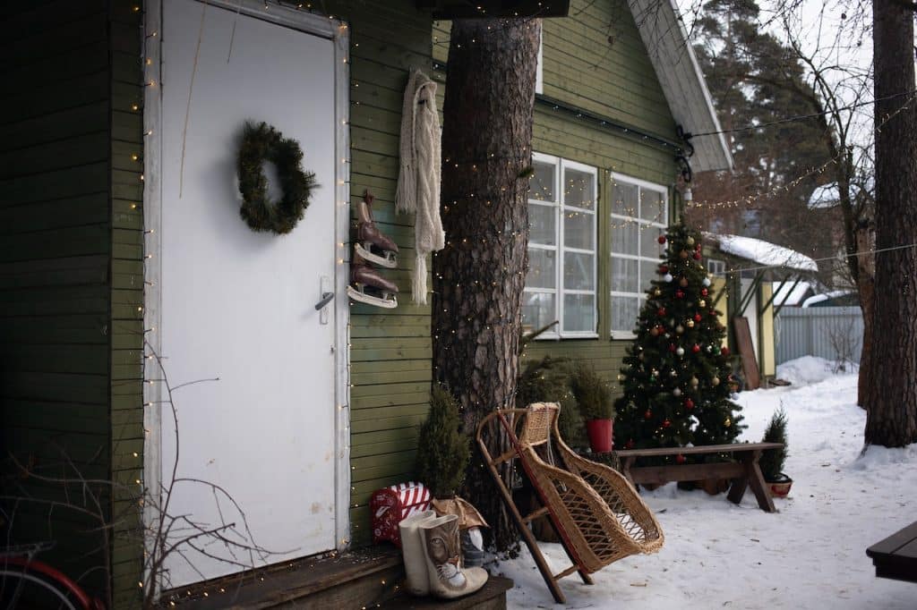 A green wooden house decorated with a holiday wreath, hanging ice skates, and a knitted scarf in a snowy setting.